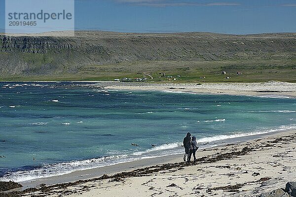 Zwei Personen spazieren entlang eines Strandes mit klarem blauen Wasser und Felsen im Hintergrund  Latrabjarg  Halbinsel Vestfirðir  Island  Europa