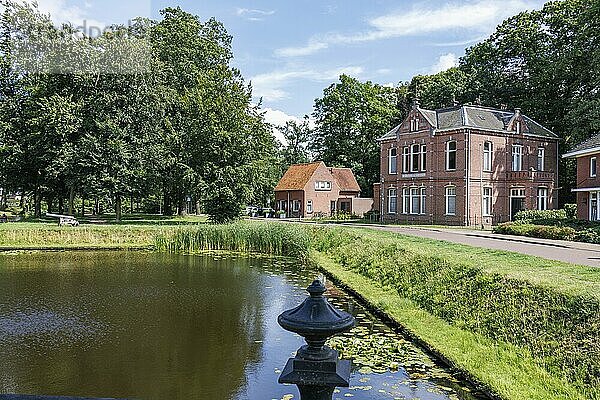 Zwei Ziegelgebäude am Flussufer in einer grünen Parklandschaft mit Bäumen und Laternen im Sommer  aalten 2