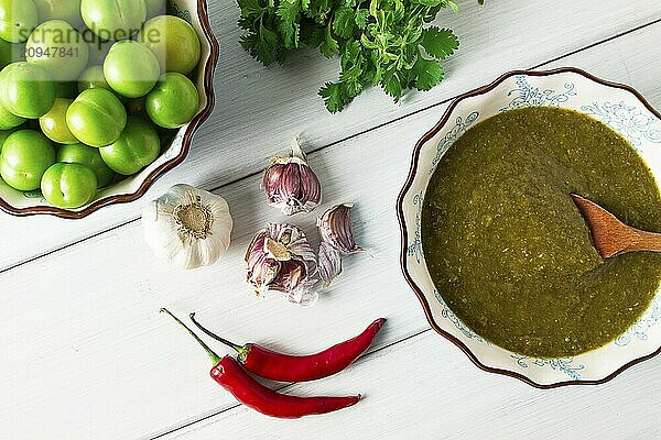 Tkemali Sauce  traditionelle georgische Küche  grüne Kirschpflaume  mit Zutaten für die Sauce  Koriander  Minze  Paprika  Knoblauch  auf einem weißen Holztisch  Nahaufnahme  rustikal  Lebensmittel Hintergrund  keine Menschen  selektiver Fokus  Sauce  grüne Sauce  tkemali  georgische Küche  hausgemacht