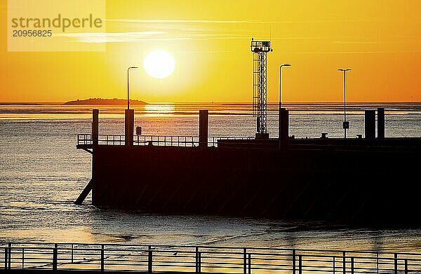 Silhouette of a jetty  pier free canal  and harbour facilities in front of a picturesque  golden  breathtakingly beautiful sunset over the sea  island Langlütjen II far away  sun ball just above the horizon  orange glowing cloudless sky  wide sea in soft colours  calm atmosphere  mouth of the Weser into the North Sea  Outer Weser  Bremerhaven  Land Bremen  Germany  Europe