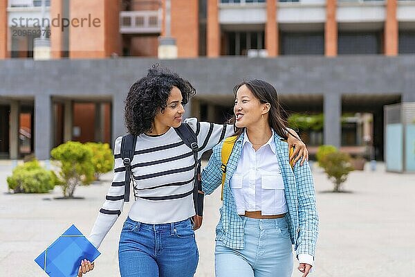 Frontal three quarter length photo of two multi-ethnic female student embracing walking along the university campus