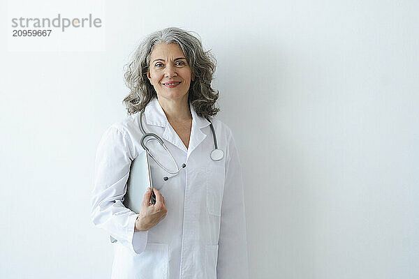 Confident senior doctor with tablet PC standing in front of white wall