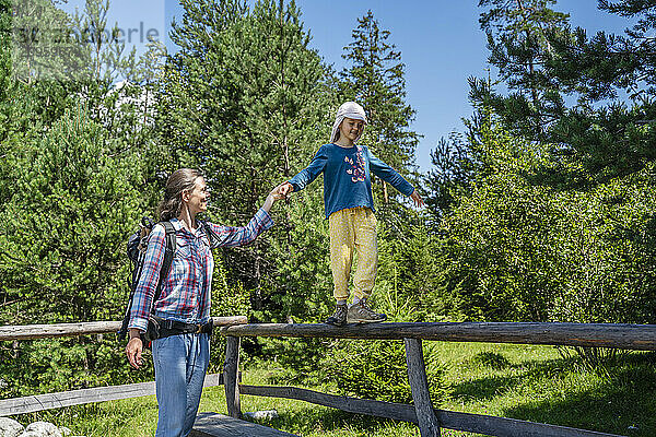 Girl balancing on fence holding mother's hand in forest