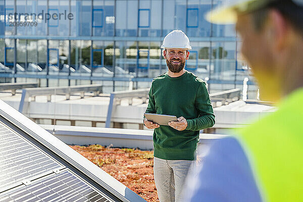 A man on the rooftop of a solar-powered building  holding a tablet with a smile on his face