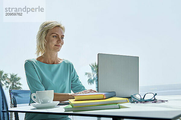 Freelance professional working on a laptop near a window with books and coffee.