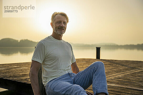 Smiling senior man sitting on edge of pier near lake at sunrise