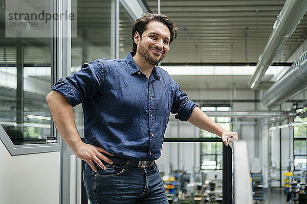 Smiling young businessman standing with hand on hip near railing in factory