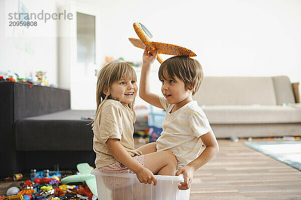 Cute boy holding airplane toy and sitting in plastic container with sister at home