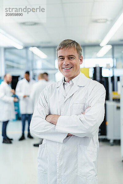 Technician in white lab coat standing in electronics factory with arms crossed