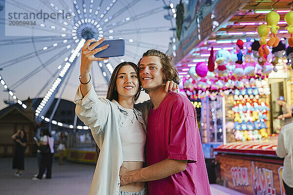 Happy couple hugging and taking selfie through smart phone at amusement park