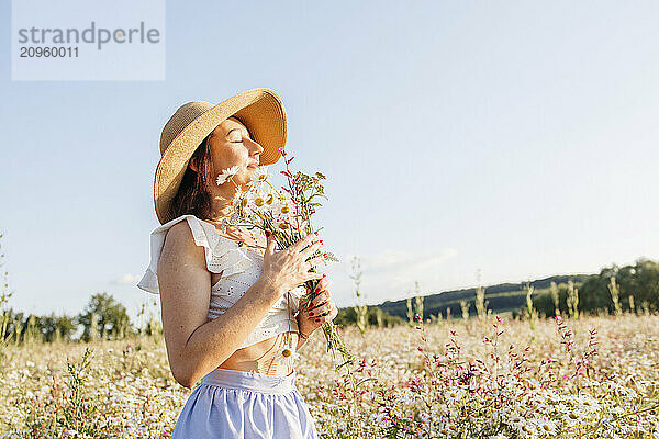 Woman wearing sun hat smelling daisy flowers in field