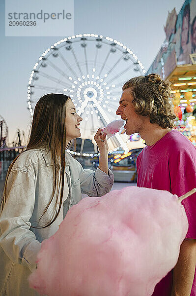 Cheerful girlfriend feeding cotton candy to boyfriend at amusement park