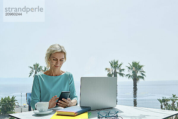 Freelance woman using smartphone while working at laptop near the window.