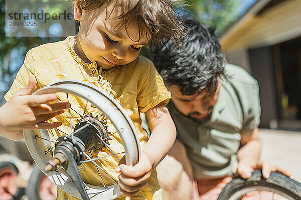 Boy holding wheel rim and repairing bicycle with father in back yard