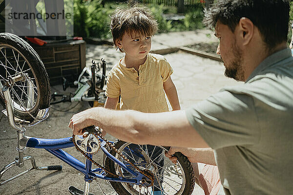 Boy standing with father repairing bicycle in back yard