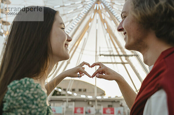 Couple making heart symbol with finger in front of ferris wheel at amusement park