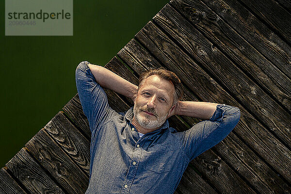 Senior man with hands behind head lying on pier near lake