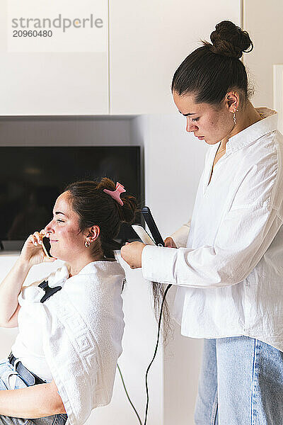 Smiling woman talking on smartphone getting hair straightened by friend at home