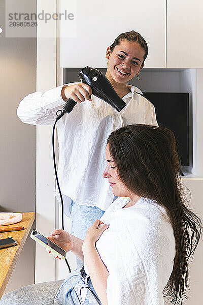 Smiling woman using hairdryer to groom friend's hair at home