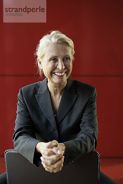 Happy businesswoman with hands clasped sitting near red wall