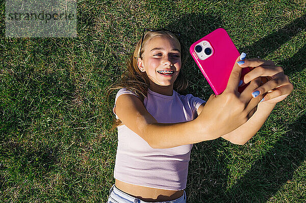 Happy girl lying down on grass and taking selfie through smart phone