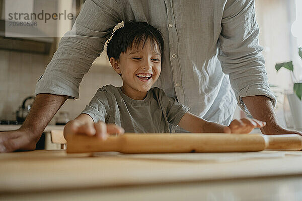 Smiling boy rolling dough with father in kitchen at home