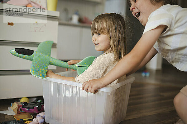 Smiling girl holding toy airplane and sitting in plastic container at home