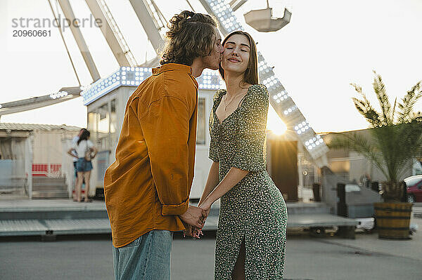 Boyfriend kissing girlfriend on cheek at sunset in amusement park