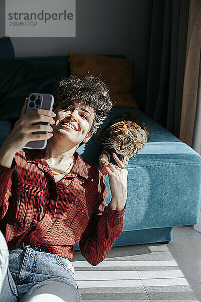 Cheerful woman taking selfie and playing with cat at home