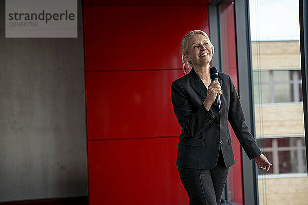 Happy senior businesswoman holding microphone near red wall