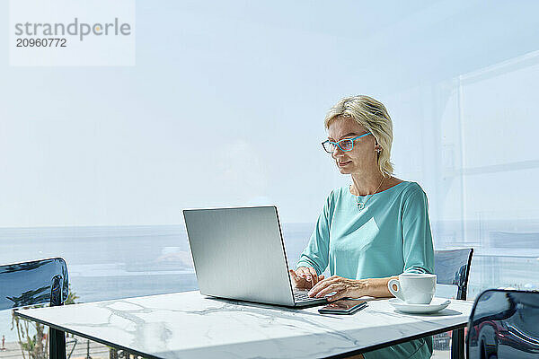 Blonde woman working on a laptop at a table indoors near a window.