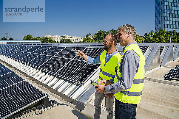 Two workers in safety vests discuss on a rooftop with solar panels while holding a tablet computer