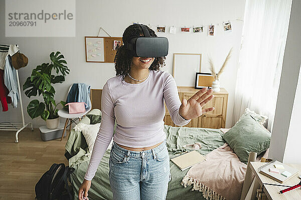 Smiling teenage girl wearing virtual reality simulators gesturing in bedroom at home