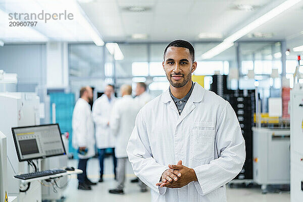 Confident technician smiling with arms crossed while colleagues are talking in electronics factory