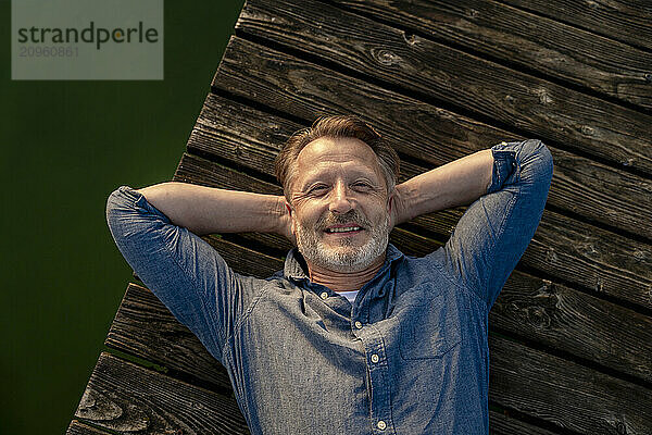 Smiling senior man with hands behind head lying on pier near lake