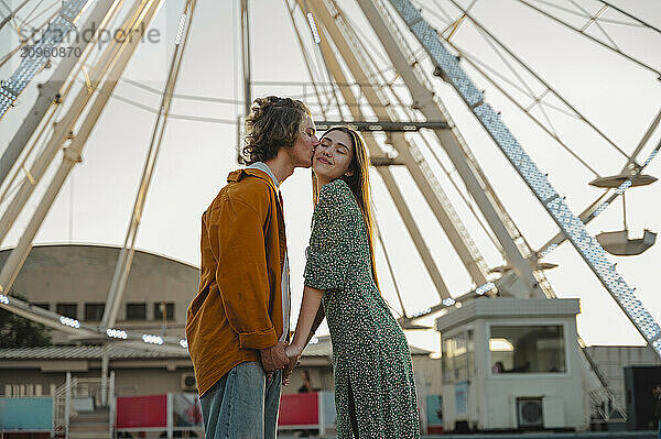 Boyfriend kissing girlfriend standing in front of ferris wheel at amusement park