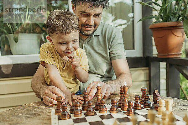 Son with hand on chin playing chess by father on porch