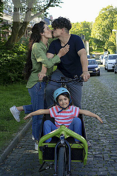 Couple kissing and standing near daughter sitting in cargo bike on cobbled street