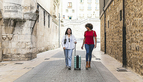 Smiling woman holding suitcase and walking with friend at street