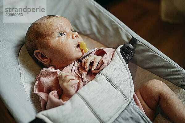 Cute newborn baby girl with pacifier lying in baby bouncer at home