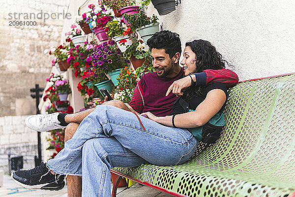 Man with arm around girlfriend using smart phone and sitting on bench