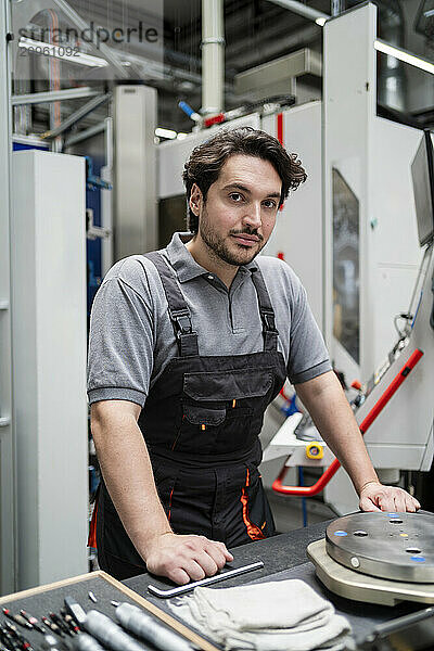 Confident young worker leaning on table with machine parts in factory