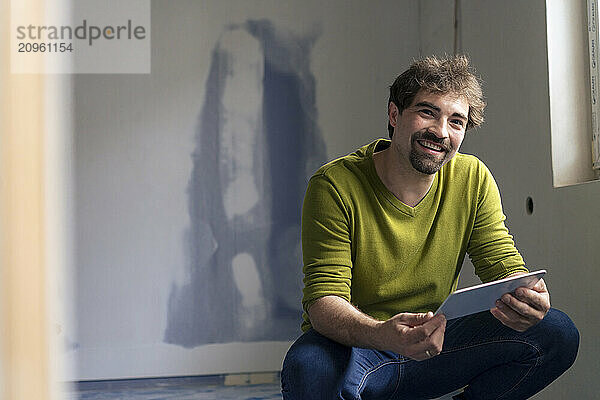 Smiling man crouching and holding tablet PC at new apartment