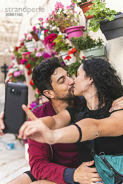 Woman kissing boyfriend and taking selfie through smart phone near potted plants