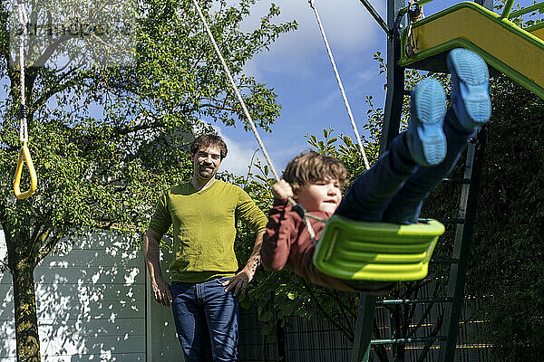 Smiling father spending leisure time with son swinging in back yard