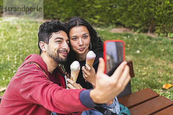 Smiling man taking selfie through smart phone and holding ice cream with girlfriend at park