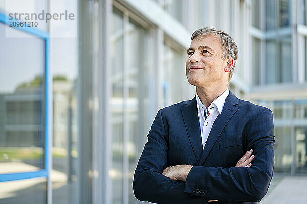 Smiling businessman standing in front of office building with crossed arms