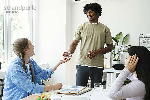 Smiling young man giving glass of water to friend sitting near table at home