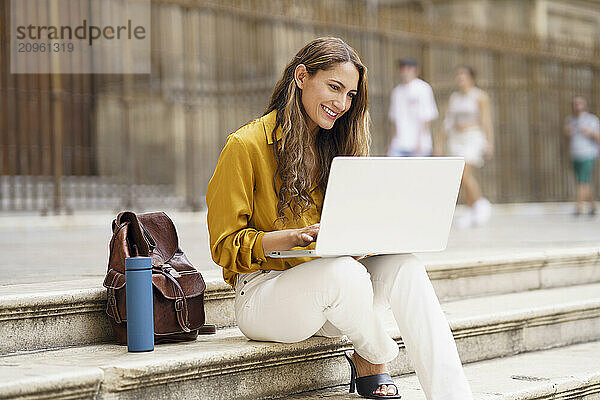 Smiling businesswoman using laptop and sitting on steps in city