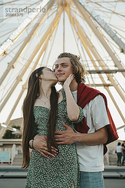 Happy girlfriend kissing boyfriend standing in front of ferris wheel at amusement park